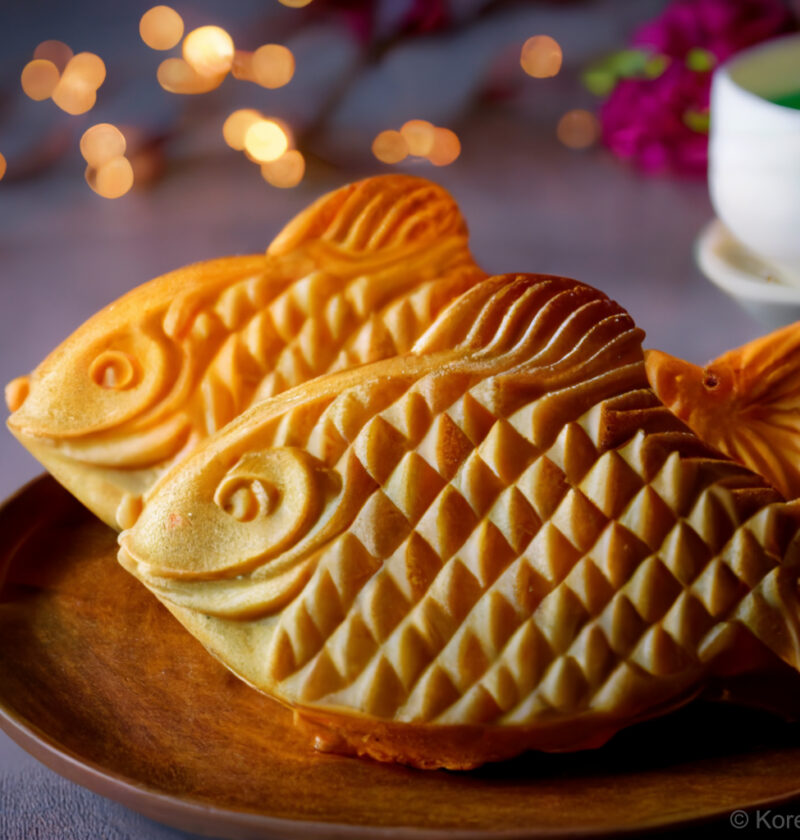 A plate of two golden, crispy Bungeoppang, a traditional Korean fish-shaped pastry filled with sweet red bean paste.